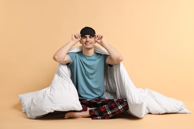 Photo of Happy man in pyjama and sleep mask wrapped in blanket on beige background