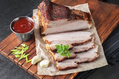 Photo of Pieces of baked pork belly served with sauce and parsley on black textured table, closeup