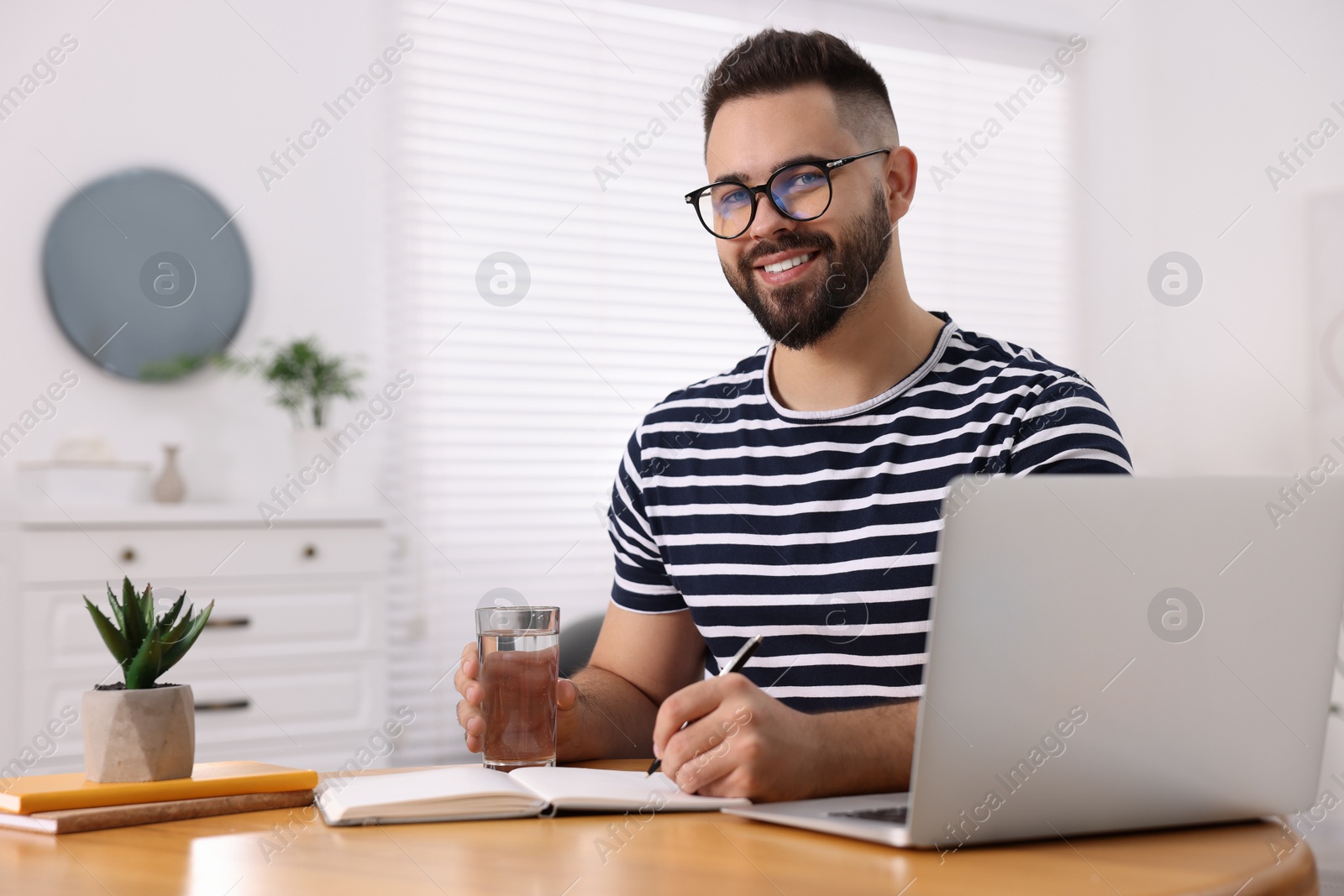 Photo of Young man with glass of water writing in notebook at wooden table indoors