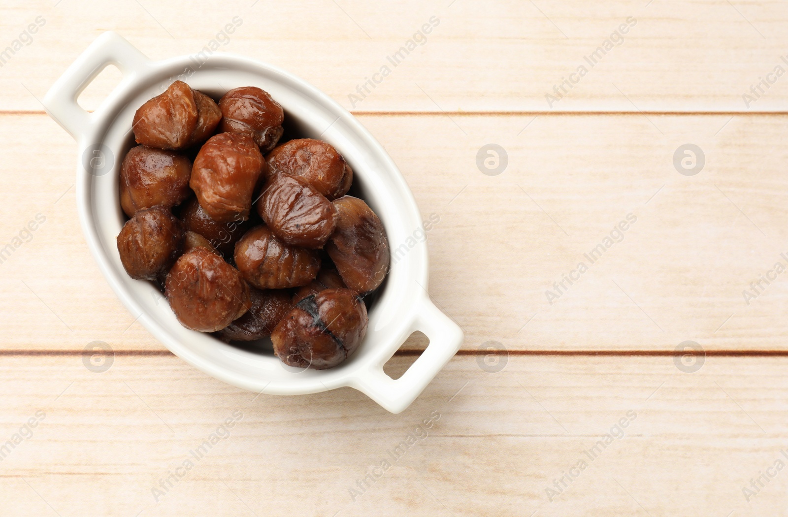 Photo of Roasted edible sweet chestnuts in dish on light wooden table, top view. Space for text