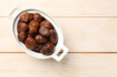 Photo of Roasted edible sweet chestnuts in dish on light wooden table, top view. Space for text