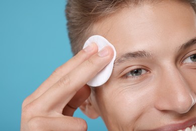 Man cleaning face with cotton pad on light blue background, closeup