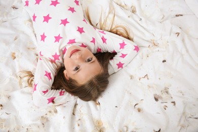 Cute little girl in pajamas lying among feathers on bed, top view. Happy childhood