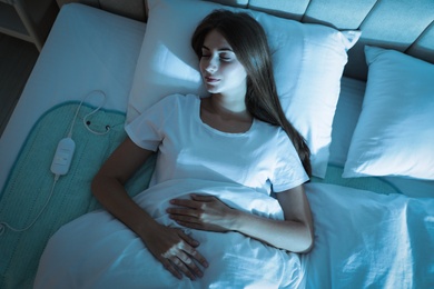 Young woman sleeping on electric heating pad in bed at night, top view