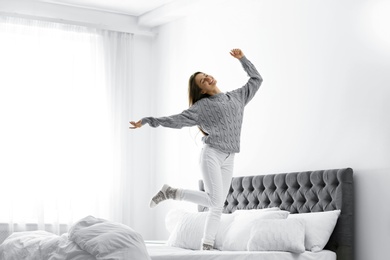 Young woman in warm sweater jumping on bed at home