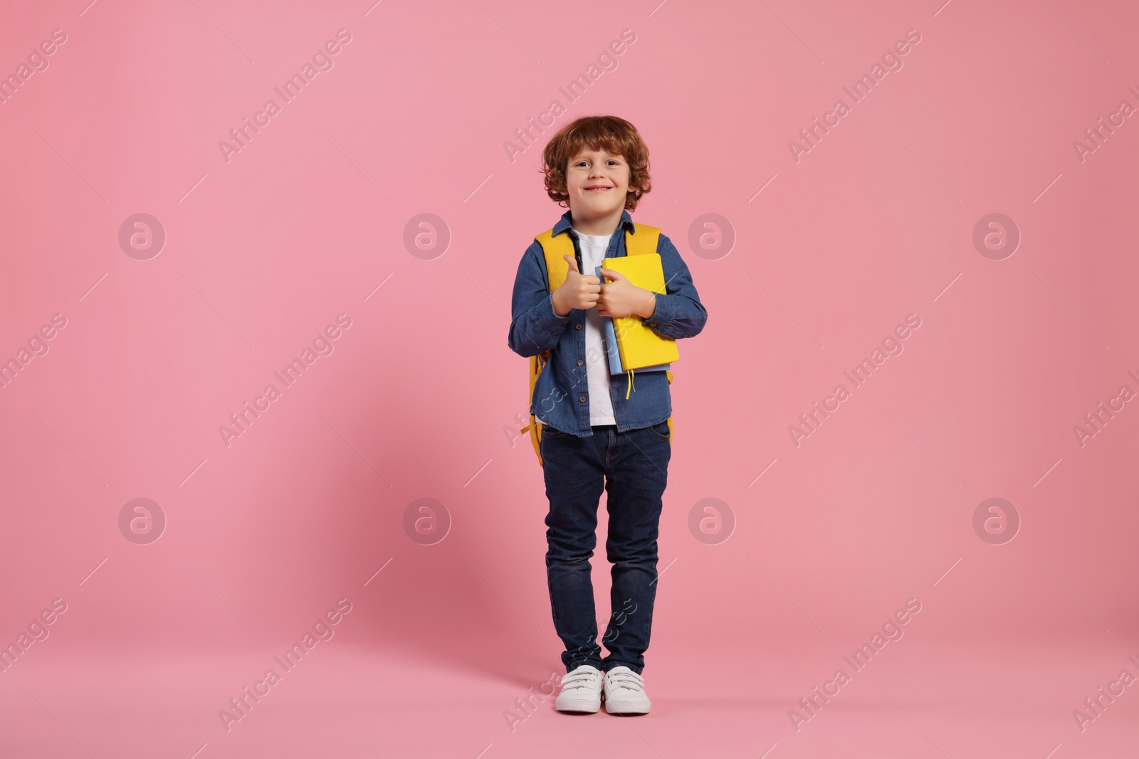 Photo of Happy schoolboy with backpack and books showing thumb up gesture on pink background