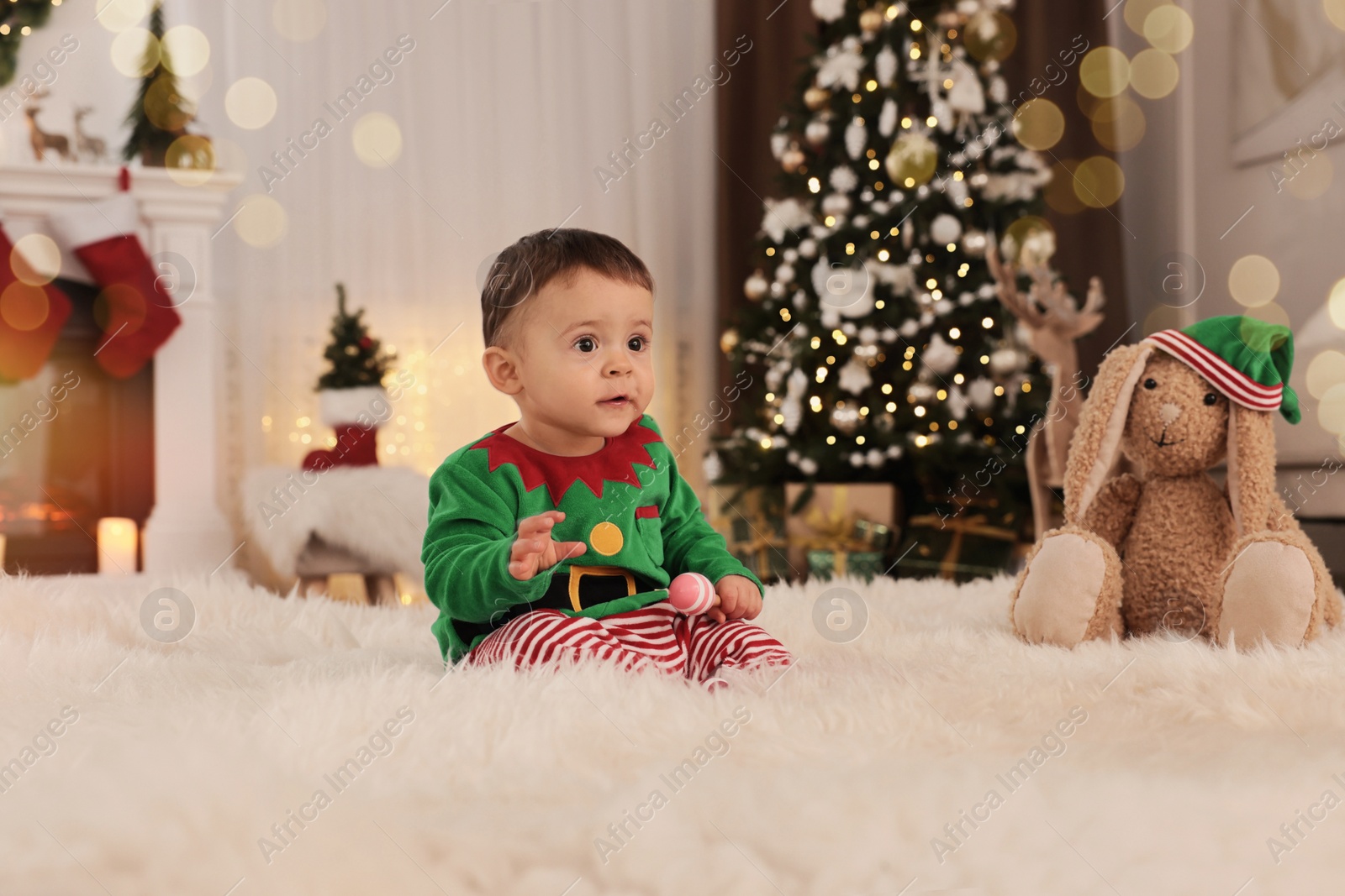 Photo of Baby wearing cute elf costume on floor in room decorated for Christmas