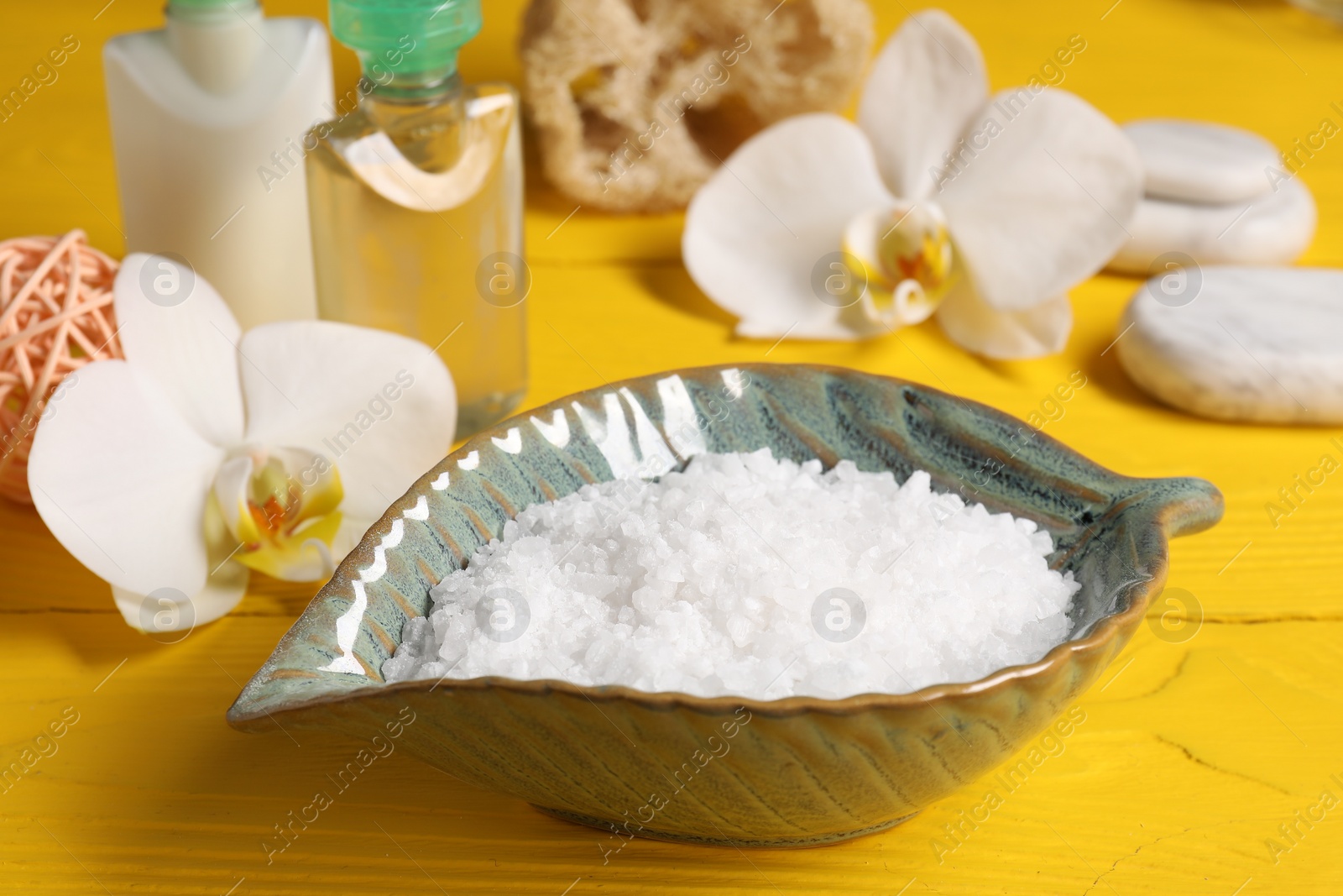 Photo of Natural sea salt in bowl and other spa products on yellow wooden table, closeup