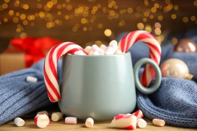 Photo of Cup of tasty cocoa with marshmallows and Christmas candy canes on wooden table against blurred festive lights