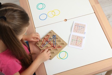 Photo of Motor skills development. Girl playing with geoboard and rubber bands at white table, above view