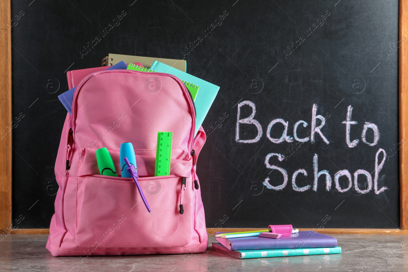 Photo of Backpack with stationery on table against blackboard with written words BACK TO SCHOOL