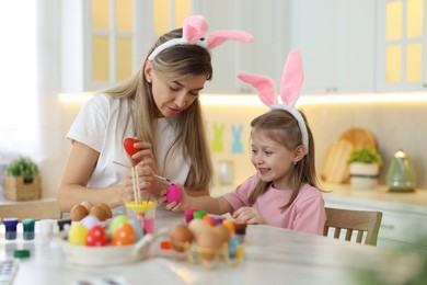 Photo of Easter celebration. Mother with her cute daughter painting eggs at white marble table in kitchen