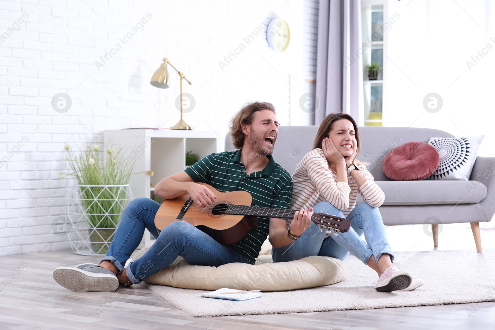 Photo of Young man playing acoustic guitar badly for displeased girlfriend in living room. Talentless musician