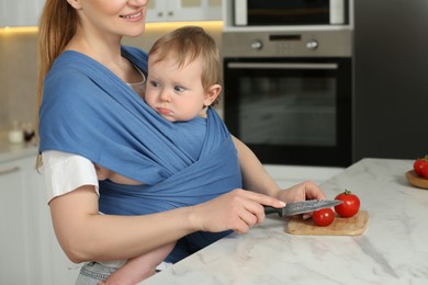 Photo of Mother cutting tomatoes while holding her child in sling (baby carrier) in kitchen