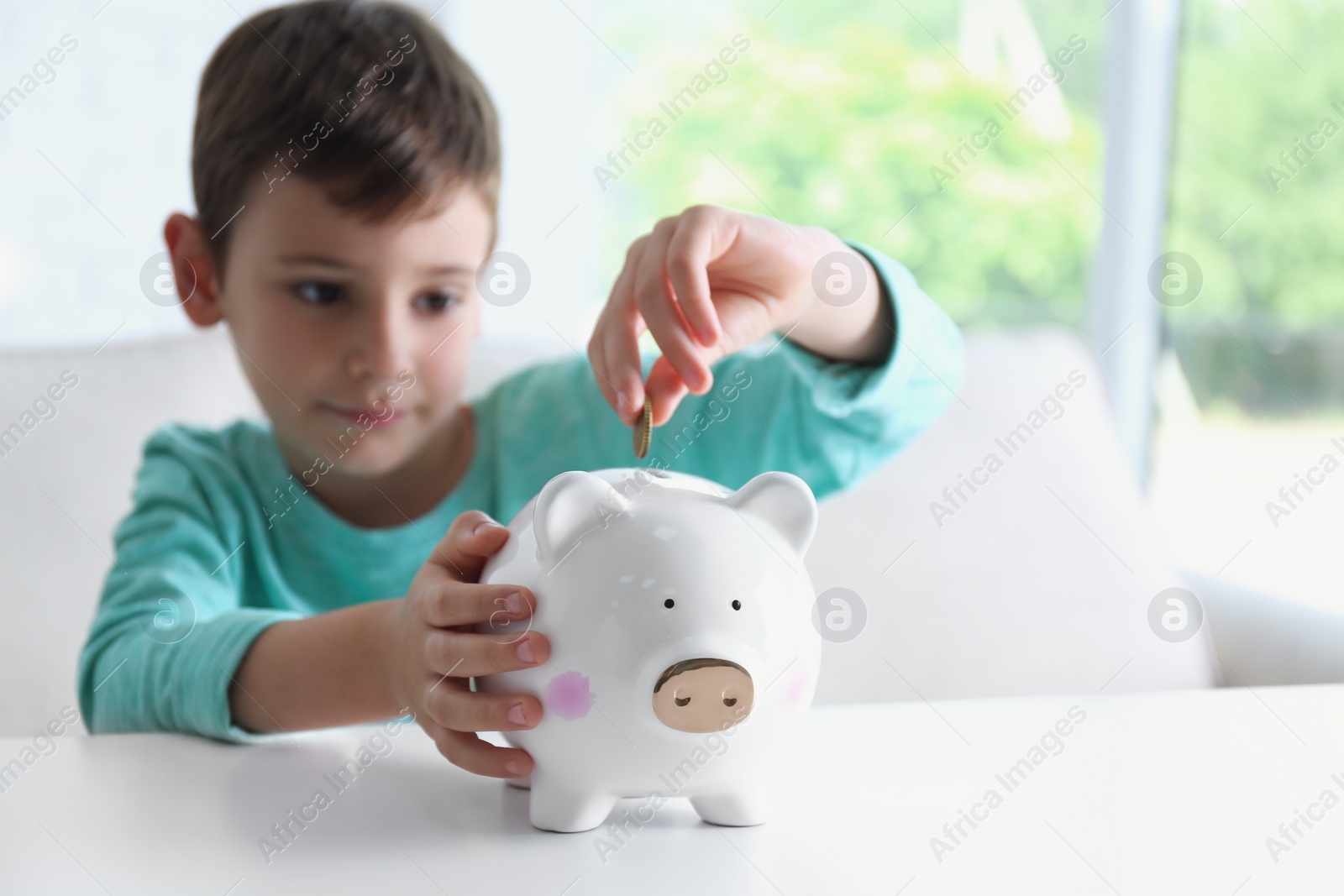 Photo of Little boy putting coin into piggy bank at white table indoors
