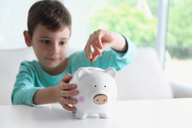 Photo of Little boy putting coin into piggy bank at white table indoors