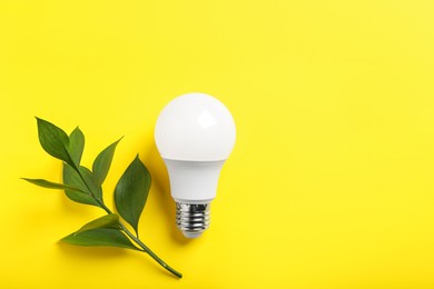 Photo of Light bulb and branch with green leaves on yellow background, flat lay. Space for text