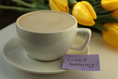 Photo of Cup of coffee, flowers and card with phrase GOOD MORNING! on white table, closeup