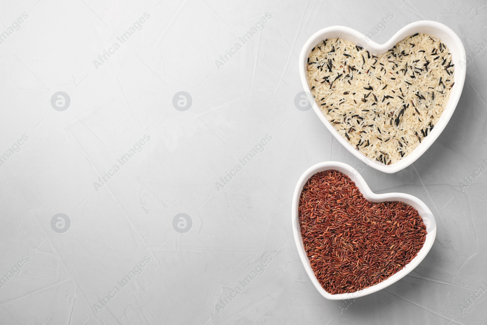 Photo of Brown and polished rice in heart shaped bowls on light grey table, flat lay. Space for text