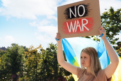 Sad woman holding poster with words No War and Ukrainian flag outdoors