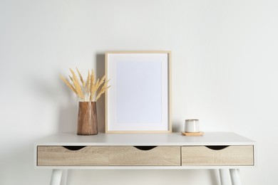 Photo of Empty photo frame, cup and vase with dry decorative spikes on white table. Mockup for design