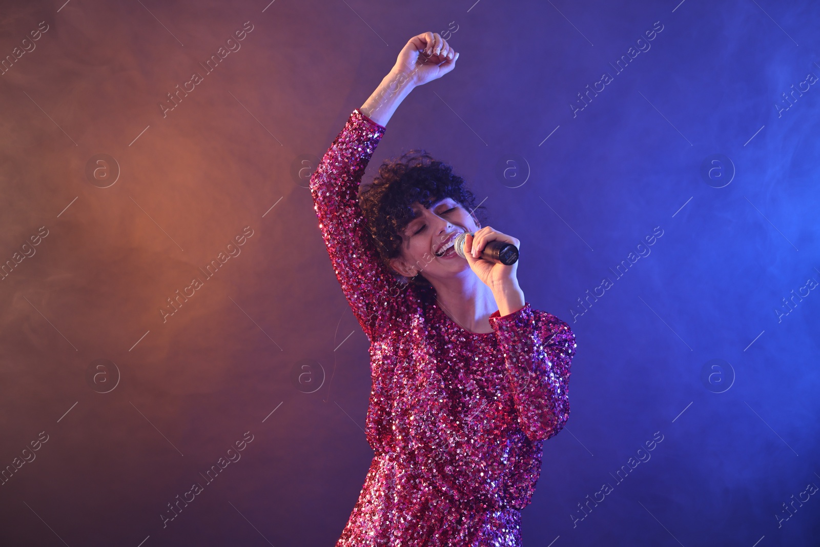 Photo of Beautiful young woman with microphone singing on color background in neon lights and smoke