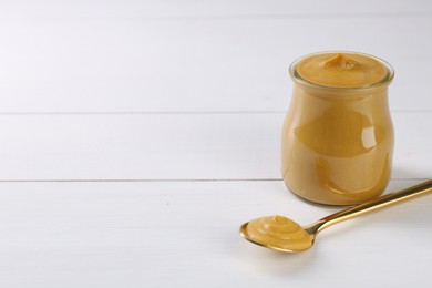 Spoon and glass jar of tasty mustard sauce on white wooden table. Space for text