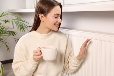 Photo of Woman holding cup with hot drink and warming hand on heating radiator indoors