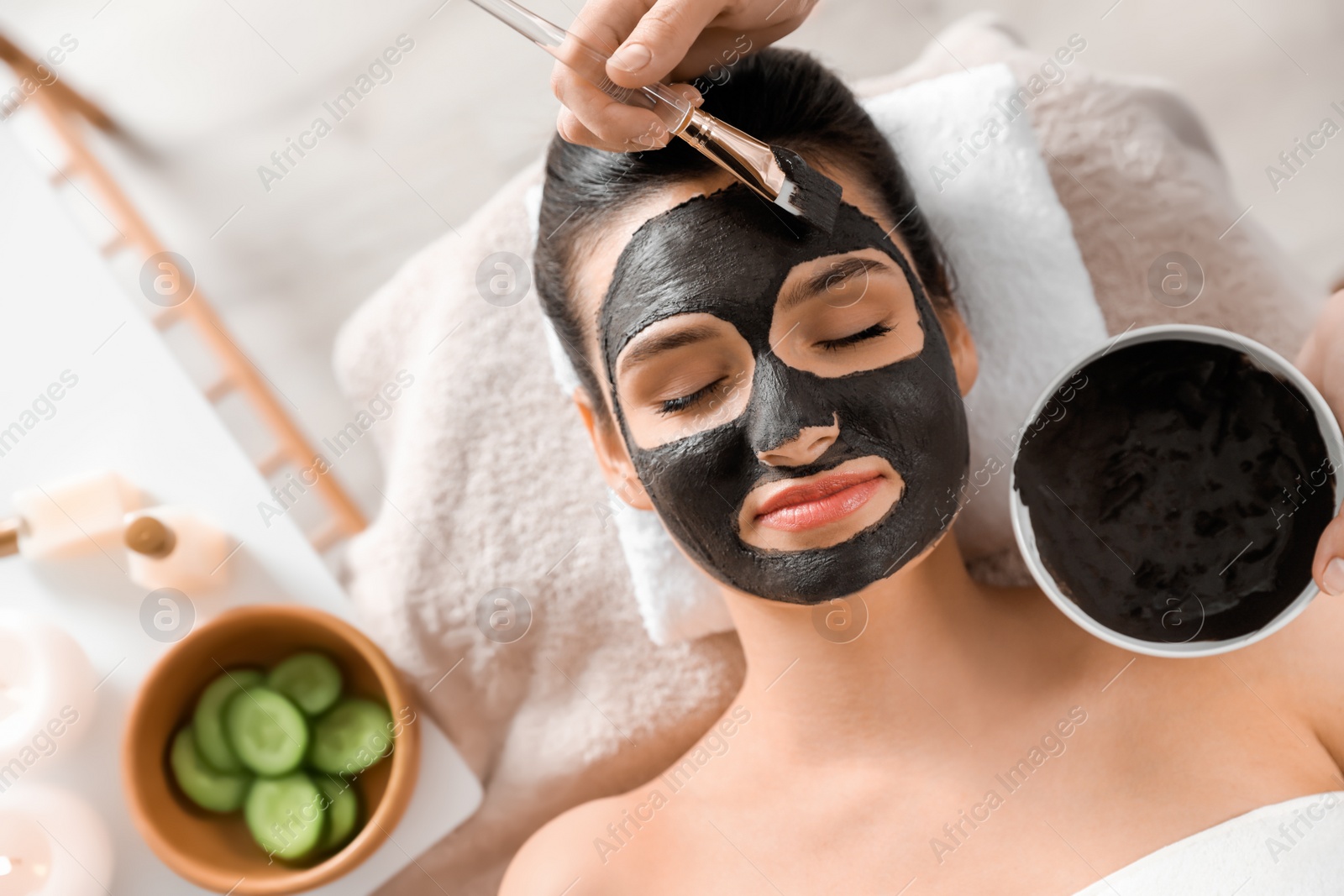 Photo of Cosmetologist applying black mask onto woman's face in spa salon, top view