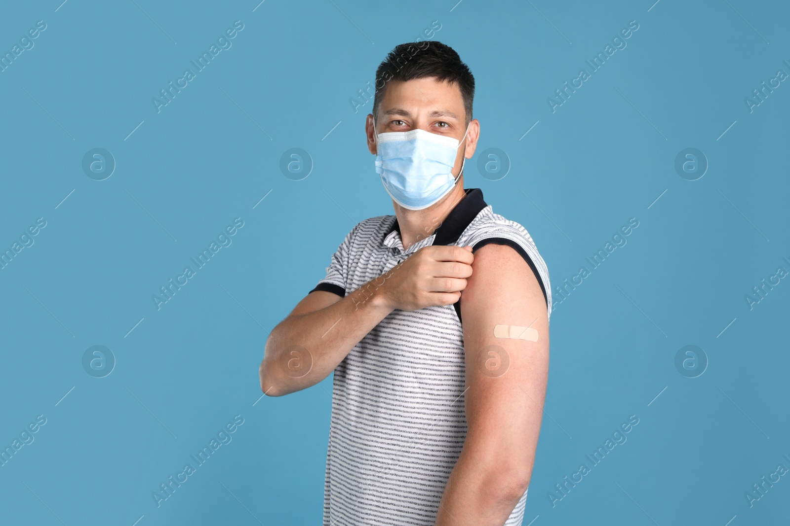 Photo of Vaccinated man with protective mask showing medical plaster on his arm against light blue background