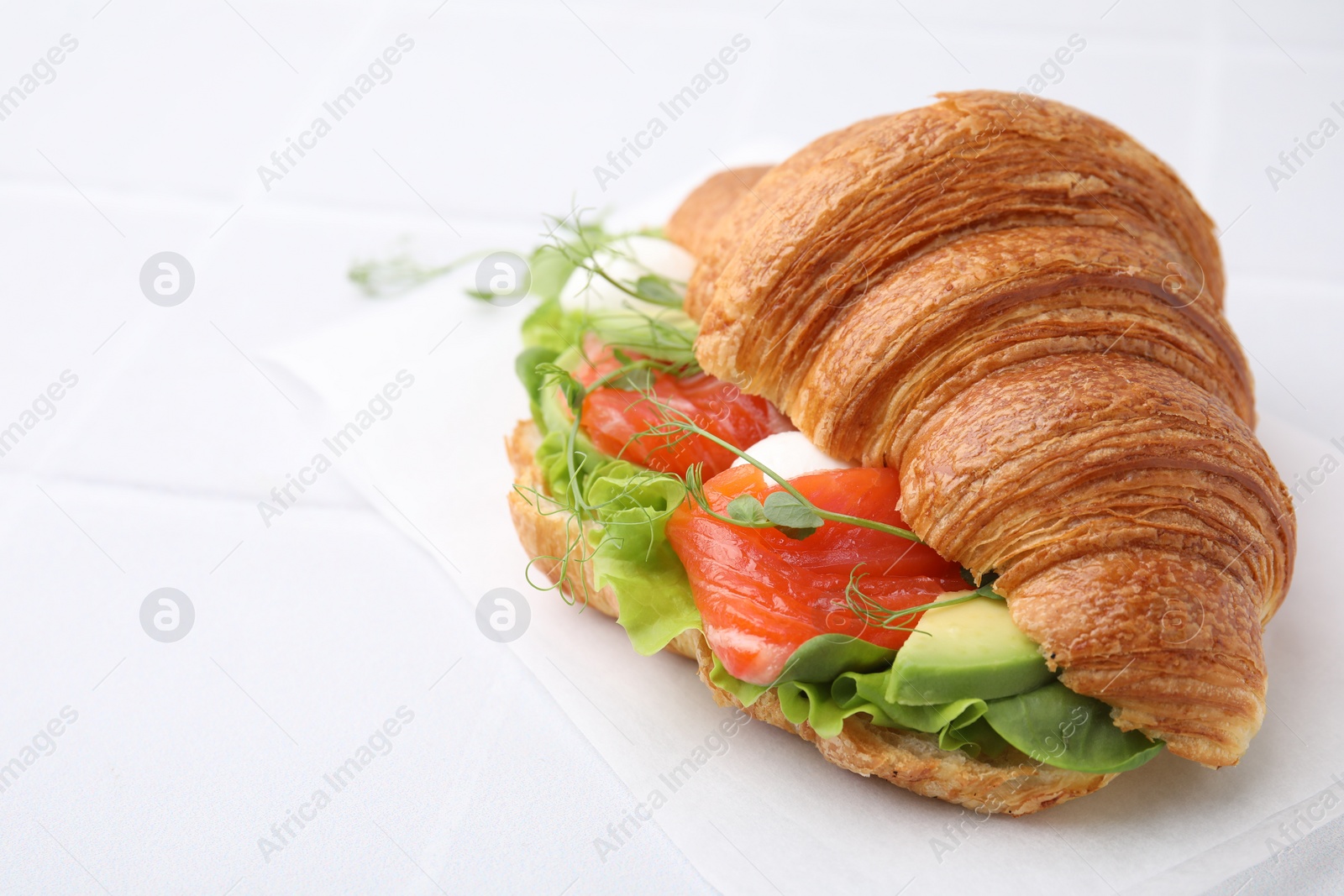 Photo of Tasty croissant with salmon, avocado, mozzarella and lettuce on white table, closeup. Space for text