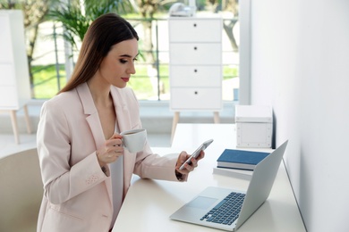Image of Young woman drinking coffee while using phone in office