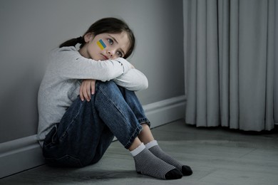 Image of Upset little girl with picture of Ukrainian flag on cheek sitting on floor indoors. Stop war in Ukraine