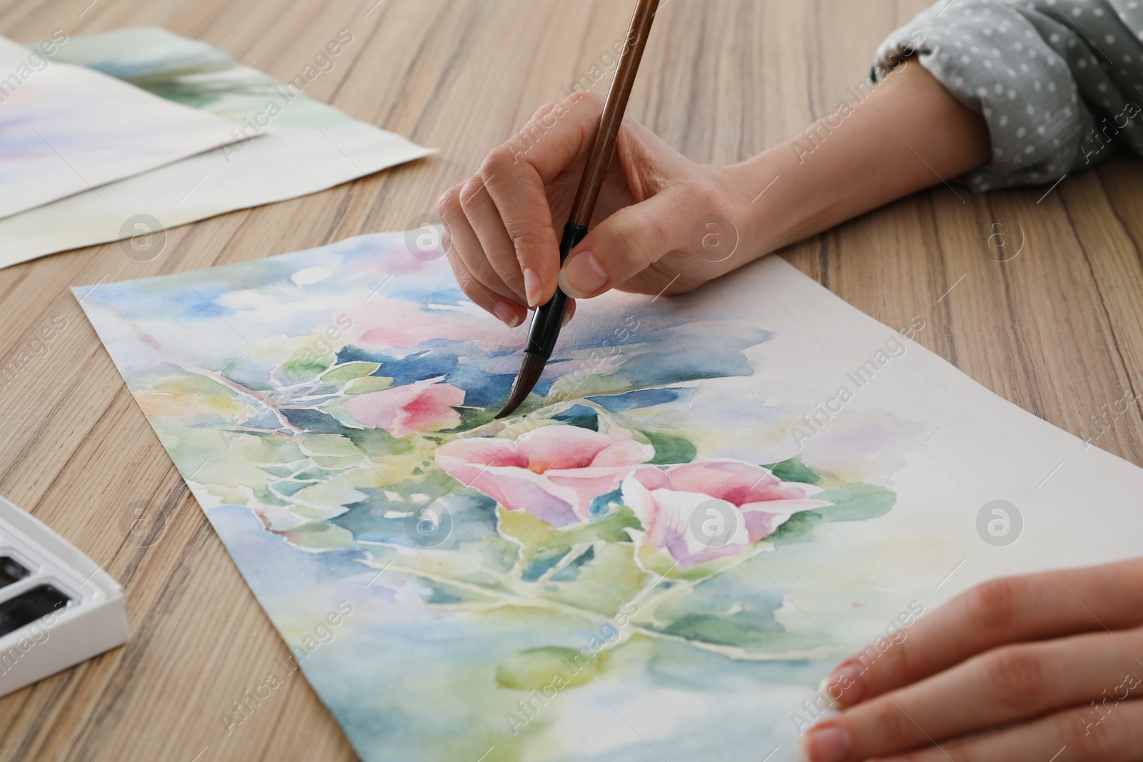 Photo of Woman painting flowers with watercolor at wooden table, closeup. Creative artwork