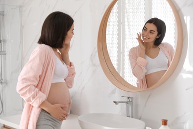 Young pregnant woman applying cosmetic product on face in bathroom