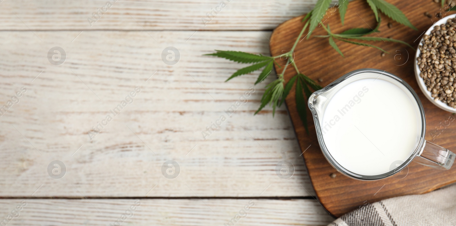 Photo of Glass jug with fresh hemp milk, seeds and leaves on white wooden table, flat lay. Space for text