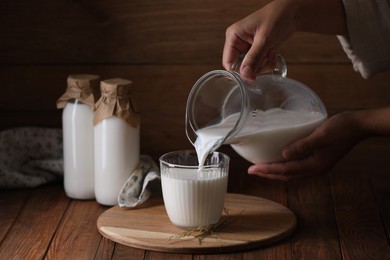 Photo of Woman pouring fresh milk from jug into glass at wooden table, closeup