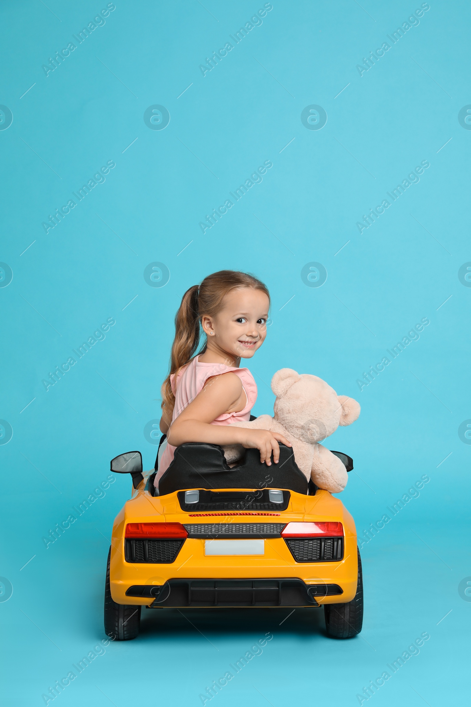 Photo of Cute little girl with toy bear driving children's car on light blue background