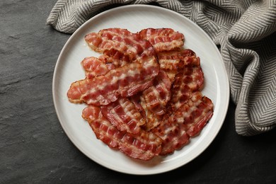 Plate with fried bacon slices on dark textured table, top view