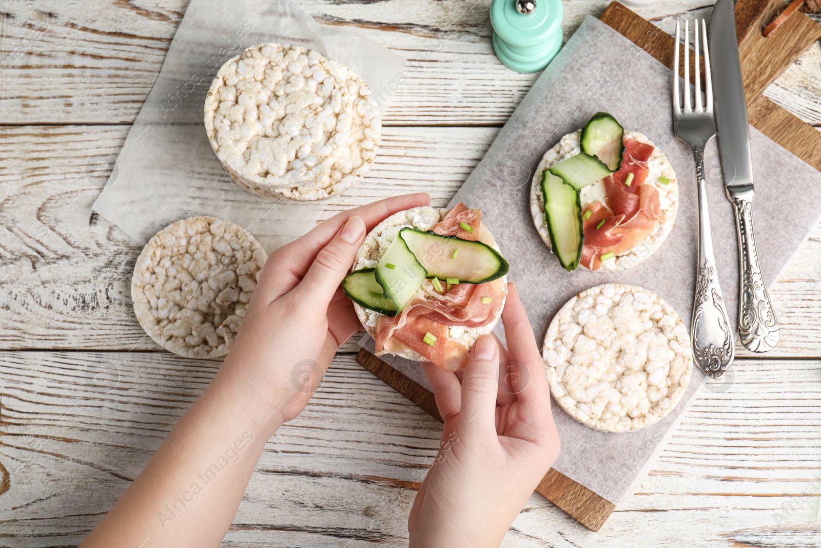 Photo of Woman holding puffed rice cake with prosciutto and cucumber at white wooden table, top view