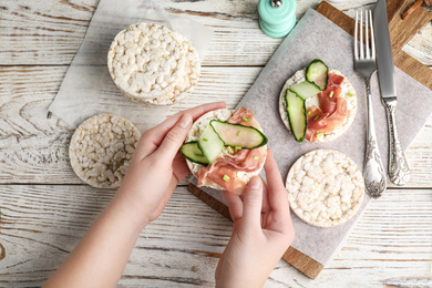 Woman holding puffed rice cake with prosciutto and cucumber at white wooden table, top view