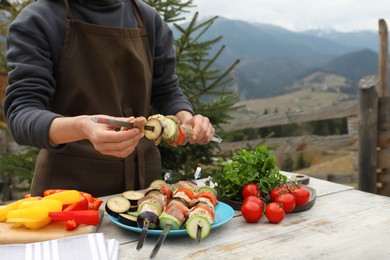 Photo of Woman stringing marinated meat and vegetables on skewer at wooden table against mountain landscape, closeup