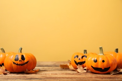 Photo of Pumpkins with scary faces and fallen leaves on wooden table against yellow background, space for text. Halloween decor