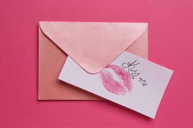 Photo of Card with lip print, phrase Kiss Me and envelope on pink background, top view