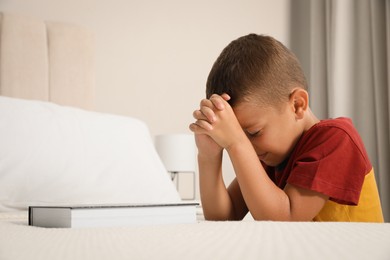 Photo of Cute little boy with hands clasped together saying bedtime prayer over Bible at home. Space for text