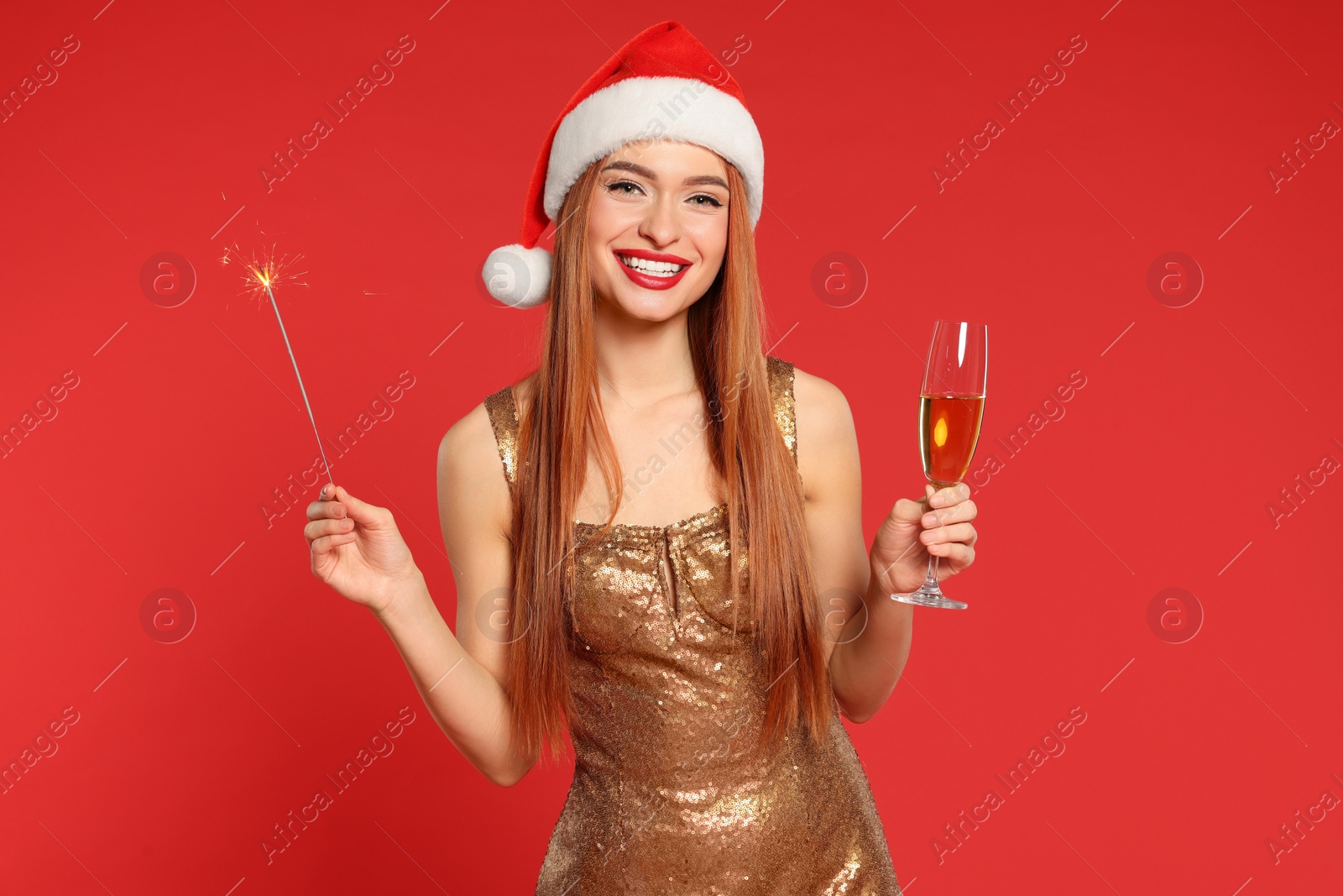Photo of Young woman in Santa hat with burning sparkler and glass of wine on red background. Christmas celebration