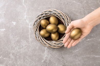 Photo of Woman holding golden egg near nest on table, top view