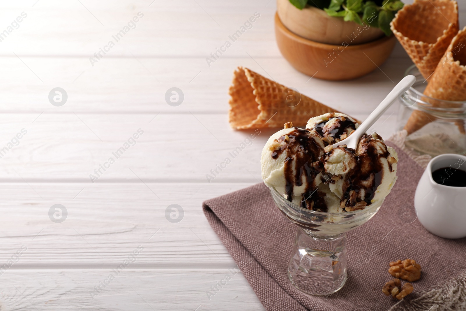 Photo of Glass dessert bowl of tasty ice cream with chocolate topping and nuts served on white wooden table. Space for text