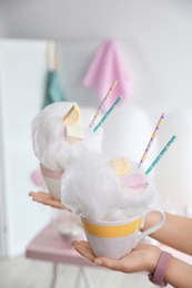 Photo of Young woman holding cups with cotton candy dessert on blurred background, closeup. Space for text