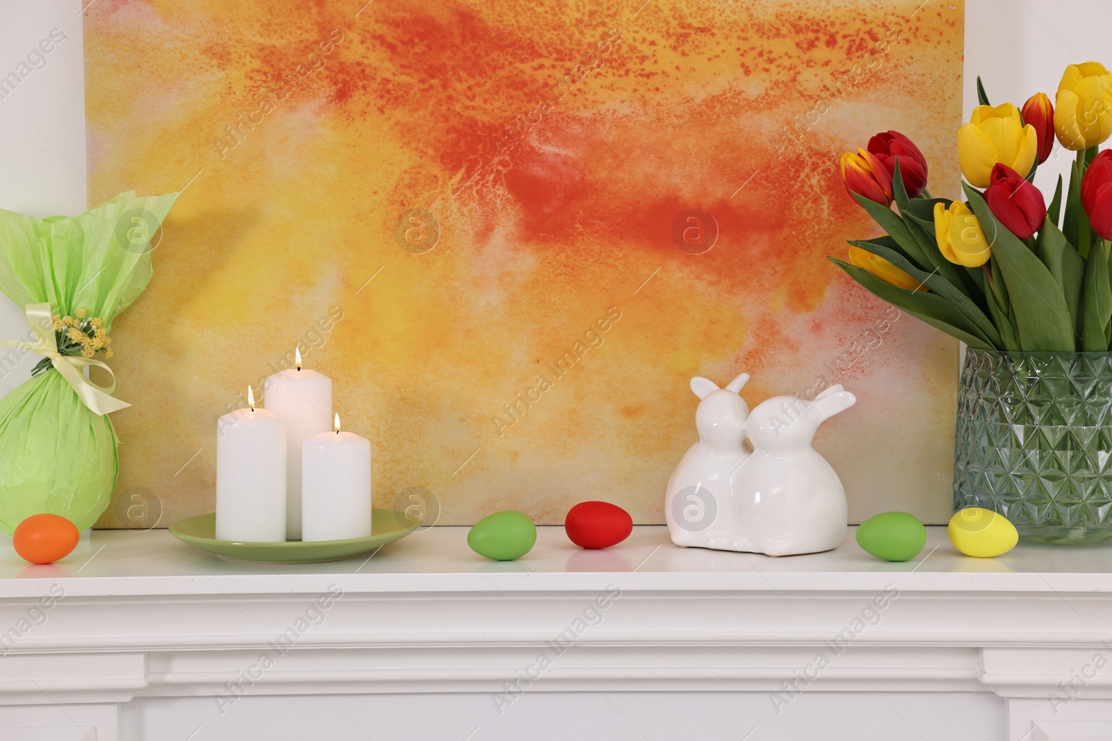 Photo of Easter decorations. Bouquet of tulips in vase, burning candles and bunny figures on fireplace at home